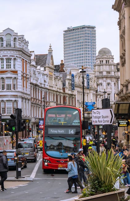 A busy urban street scene in Putney showing a red double-decker bus with the destination sign reading 'Putney Heath' parked at a bus stop amidst heavy traffic and pedestrians crossing the road. The background includes a mix of historic and modern buildings, with ornate white facades, detailed architectural features, and a contemporary high-rise building further behind. On the right side, there is a small outdoor café with black signage and a yellow sign for tobacco products, with groups of people gathered nearby. A street sign directs to The City, Trafalgar Square, and Hyde Park Corner, while another points to the Trafalgar Square area. The scene is captured under overcast daylight, bringing out a muted color palette that highlights the busy, structured environment typical of East Putney. This visual context emphasizes the importance of efficient rubbish clearance and private waste handling, as services like Waste Clearance Putney often manage waste removal from such crowded and dense urban areas, especially in estate and flat complexes.