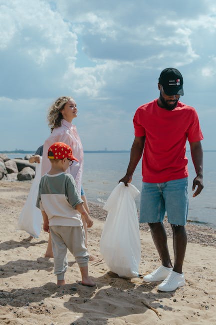 A man with dark skin, wearing a red t-shirt, distressed denim shorts, white sneakers, and a black cap with red, white, and black stripes, is standing on a sandy beach near the water's edge. He is holding a white plastic bag and appears to be in the process of collecting or disposing of waste. Behind him, a woman with light skin and curly blonde hair, dressed in a light pink jacket, and two children—one with light skin, wearing a grey t-shirt, beige shorts, and a red cap with yellow and white patterns, and the other with light skin, in a grey shirt with white and pink accents and khaki pants—are engaged in a litter collection activity. The background features rocky shoreline, calm water, and a sky filled with large fluffy clouds, typical of a bright day. The scene emphasizes outdoor environmental cleanup, dei alternative waste handling, and private rubbish removal efforts, with the boat-free waterfront setting under natural daylight and casual clothing suitable for outdoor activities.