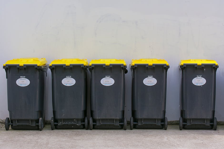 A row of five large wheeled rubbish bins positioned against a plain white wall in an indoor or sheltered area. The bins are made of dark grey plastic with textured surfaces, each fitted with yellow lids that are securely closed. White identification labels with black text are affixed to the front of each bin, displaying unique identification numbers. The bins are arranged neatly side by side on a concrete floor, with the first two on the left slightly closer to the camera and the remaining three extending further into the background. The environment appears clean and utilitarian, suitable for private waste collection or alternative rubbish removal services provided by Waste Clearance Putney. The scene emphasizes organized waste storage, ready for collection, consistent with professional rubbish removal practices, without any visible contents or external tools in the frame.