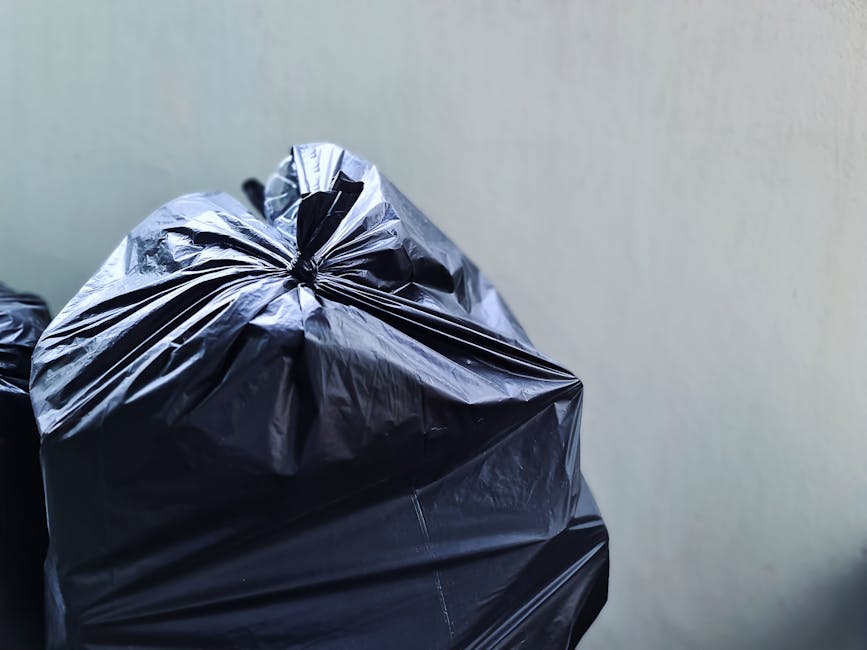 Close-up view of several tightly tied black plastic rubbish bags, stacked against a plain, light-colored wall. The bags are made of glossy, durable plastic material with visible wrinkles and creases, indicating they are filled and sealed. The primary focus is on the large bag in the foreground, which occupies most of the image space, with parts of additional bags stacked behind it. The scene is well-lit with soft, natural or diffuse light, highlighting the reflective surface of the plastic and the textures of the bags. This setting suggests an outdoor or internal waste collection point, typical for private rubbish removal services such as those provided by Waste Clearance Putney, supporting alternative waste disposal or on-site clearance activities within a domestic or commercial environment. The orderly stacking and sealing of the bags reflect a managed approach to waste collection, aligning with professional rubbish removal practices common in Putney and surrounding areas.
